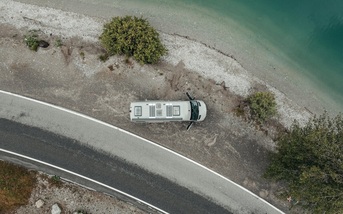 Grijze camper van bovenaf gezien, die op grind staat naast een meer aan de oever, naast het grind is er een kleine weg en op het grindbed staan drie groene bomen, de deuren van de camper zijn open