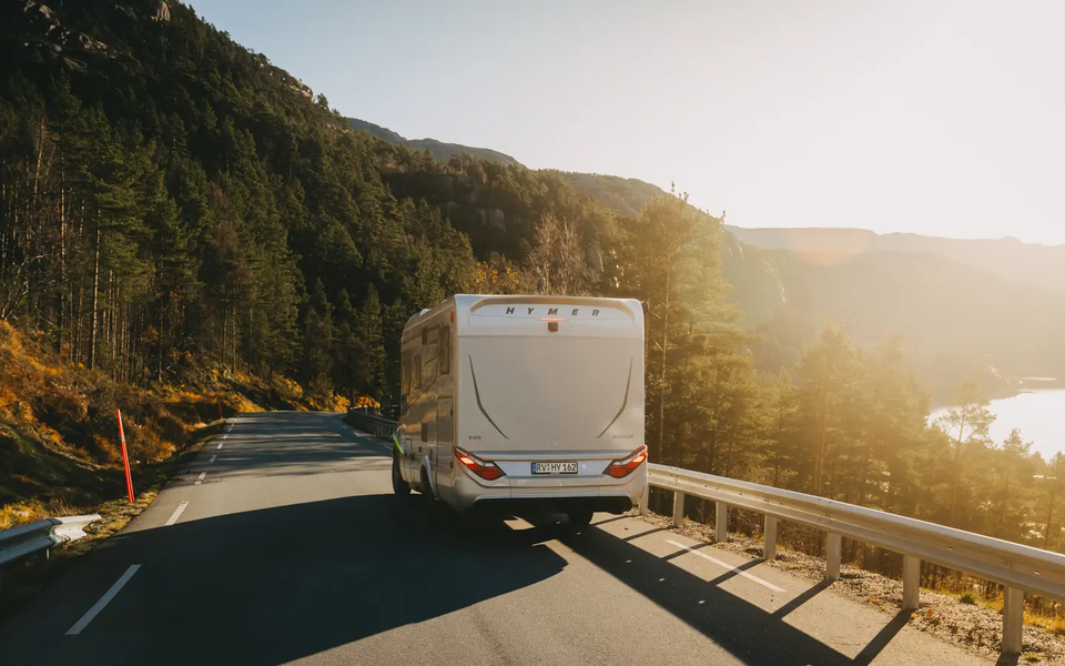 Vue arrière du HYMER B-MC T BlackLine, véhicule roulant sur une route de campagne, forêt en arrière-plan à gauche, lac en arrière-plan à droite